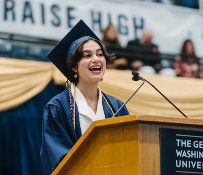 Anna Shah speaking in front of a podium with the George Washington University logo. She and those in the audience wear graduation regalia.