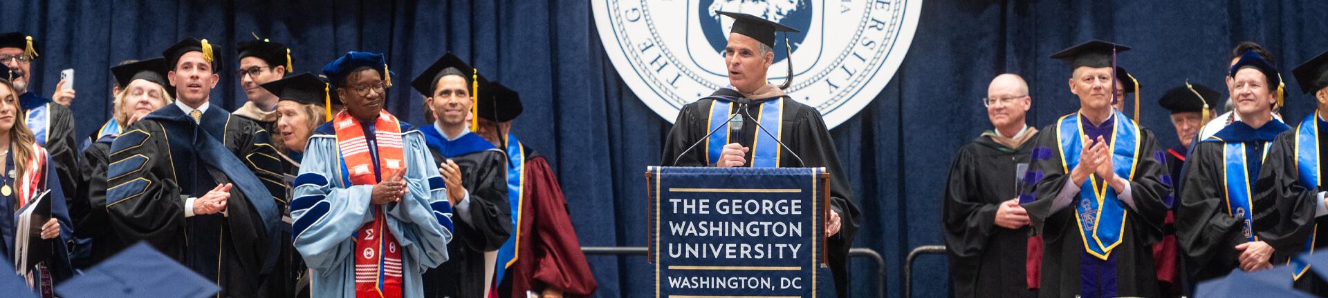 Commencement stage features speaker at podium in the center, faculty standing and clapping to the left and right